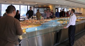 Students and candidates stand around the miniature port model while a presenter speaks.